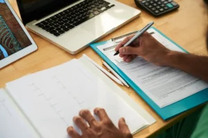Em uma mesa de trabalho, uma pessoa preenche documentos em um formulário impresso ao lado de um notebook e um celular, representando a Inscrição Online: Vantagens De Substituir Formulários Em Papel Por Sistemas Digitais.